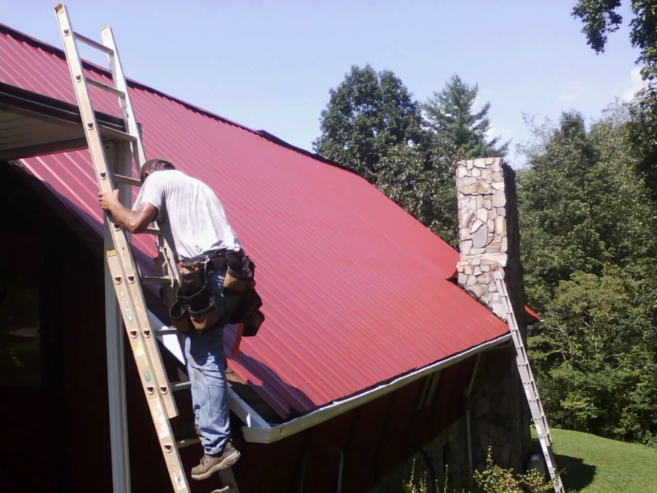 Roofer on a ladder beside a red metal roof and stone chimney — Soffit & Fascia Repair by Roofing Brothers in Crandon
